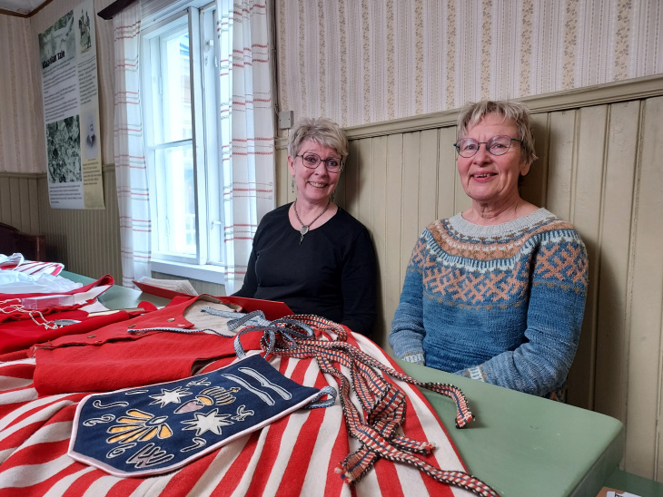 Tuusulan puvun osia pöydällä, Marjo Vainio ja Kirsi Kostamo istuu pöydän ääressä hymyillen